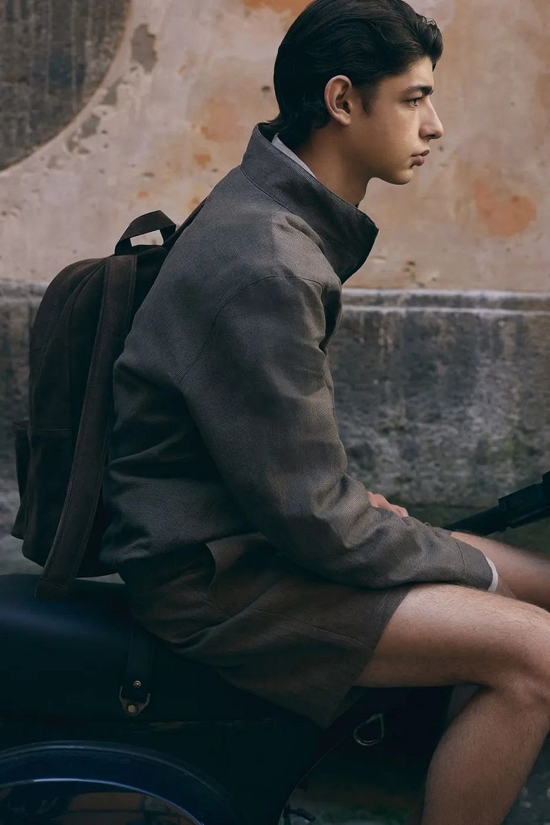 Side profile of a young man in a grey collared jacket with a brown backpack seated on a moped against a stone wall.