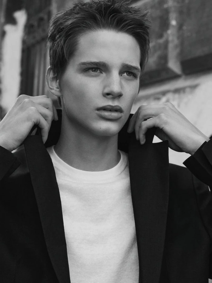 Black and white portrait of a young man adjusting the collar of his dark jacket over a white t-shirt outdoors.