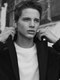Black and white portrait of a young man adjusting the collar of his dark jacket over a white t-shirt outdoors.