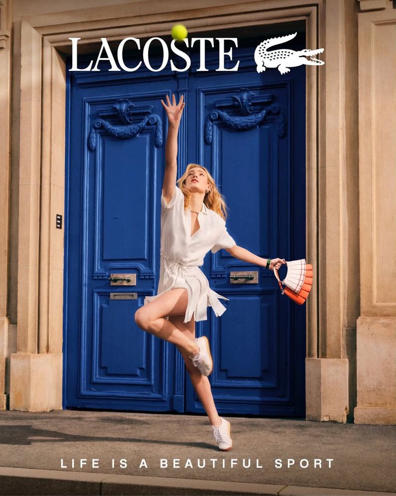 Model in white dress leaping in front of large blue double doors holding a tennis bag beneath a tennis ball