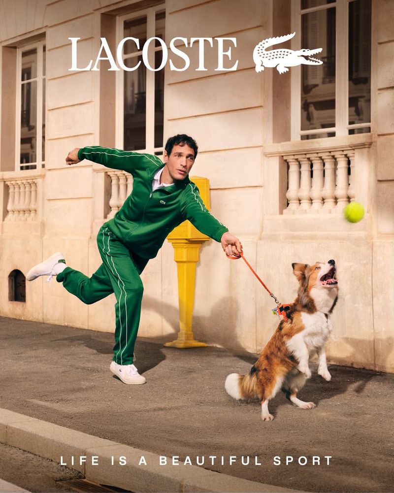Man in a green tracksuit running down a city sidewalk holding a leash as a dog jumps for a tennis ball in a Lacoste ad.