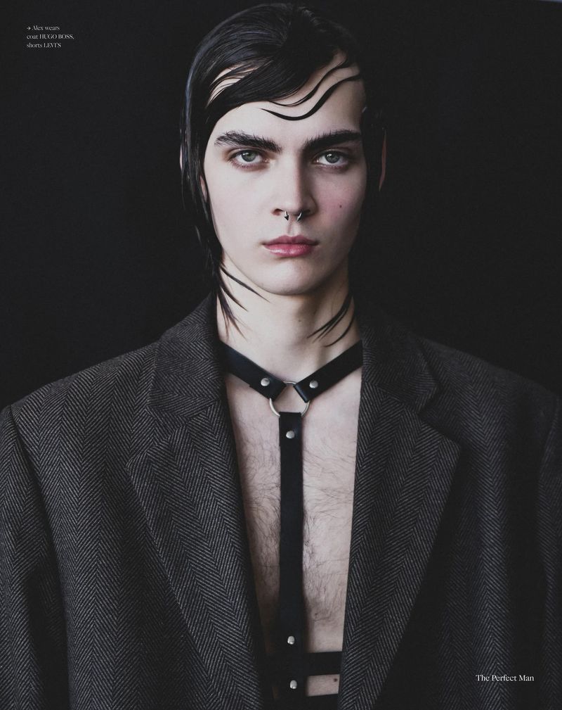 A young man with wet sculpted hair and a septum piercing wearing an open tweed blazer and leather harness against a black backdrop.