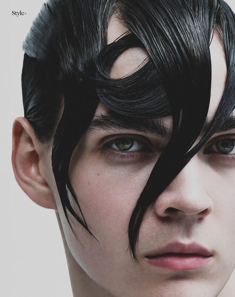 Close-up portrait of a young man with sculpted wet-look black hair draped over his forehead against a pale studio backdrop.