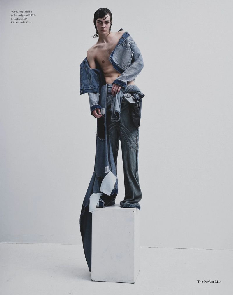 Male fashion model posing on a white plinth wearing deconstructed denim outerwear against a minimal studio backdrop.