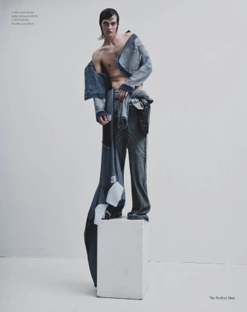 Male fashion model posing on a white plinth wearing deconstructed denim outerwear against a minimal studio backdrop.