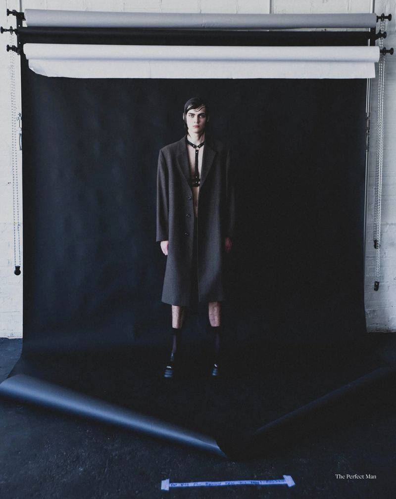 Fashion model wearing an oversized dark coat and black chest harness standing against a studio backdrop.