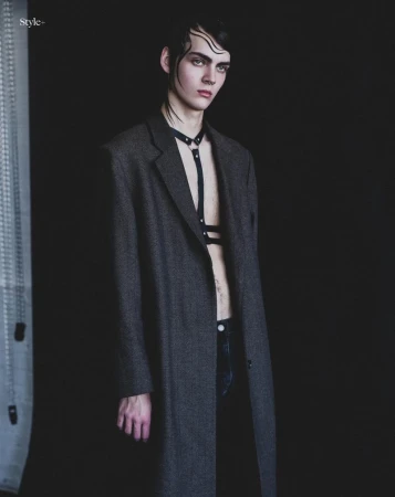 Male model wearing a long charcoal coat and black chest harness against a black studio background.