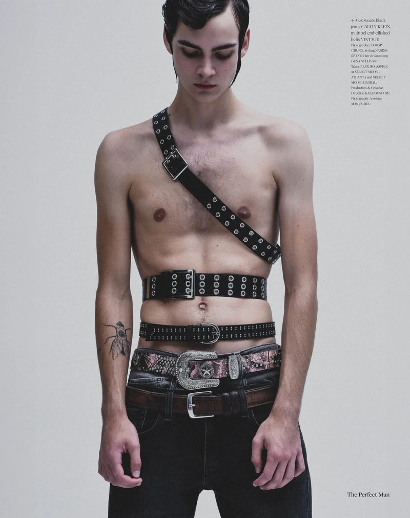 Shirtless male model wearing multiple black studded and western belts across his chest and waist against a grey studio backdrop.