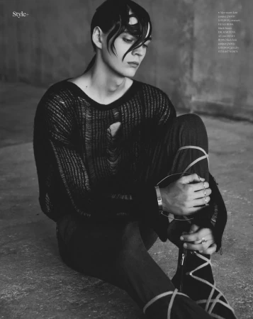 Editorial portrait of a man seated on concrete wearing a distressed black knit sweater and strapped trousers.