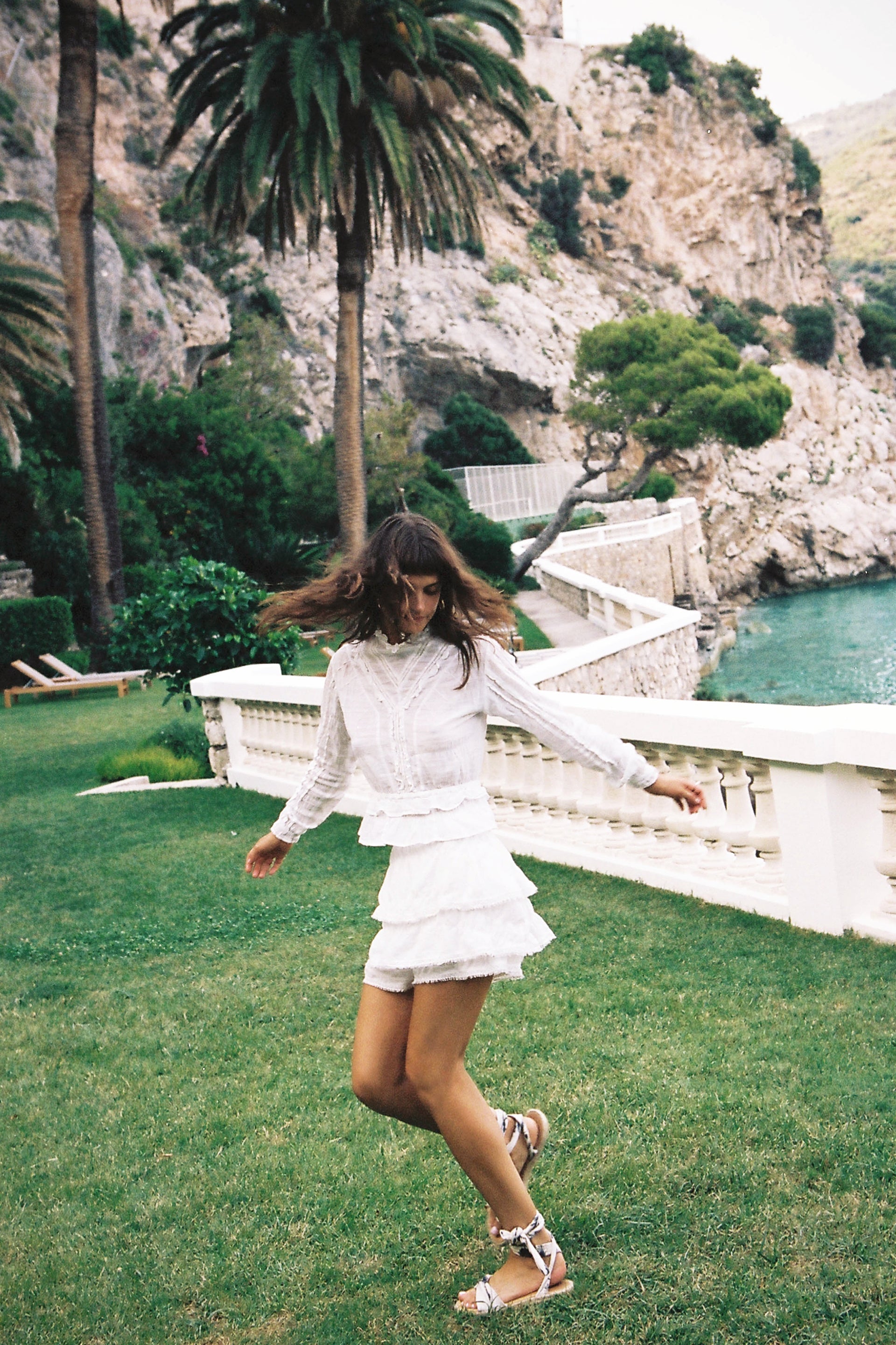 A woman in a white layered mini-skirt and sandals is dancing on a lawn in front of a cliffside with a palm tree and ocean view.