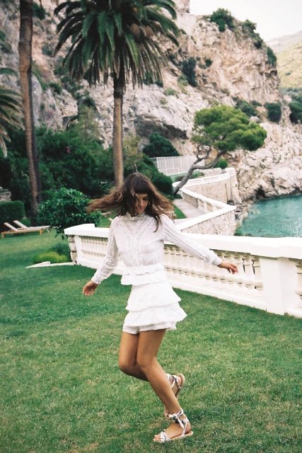 A woman in a white layered mini-skirt and sandals is dancing on a lawn in front of a cliffside with a palm tree and ocean view.