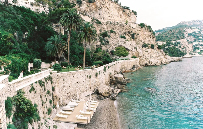 High-angle view of a woman in a white dress standing on a coastal terrace with palm trees beside the blue ocean.