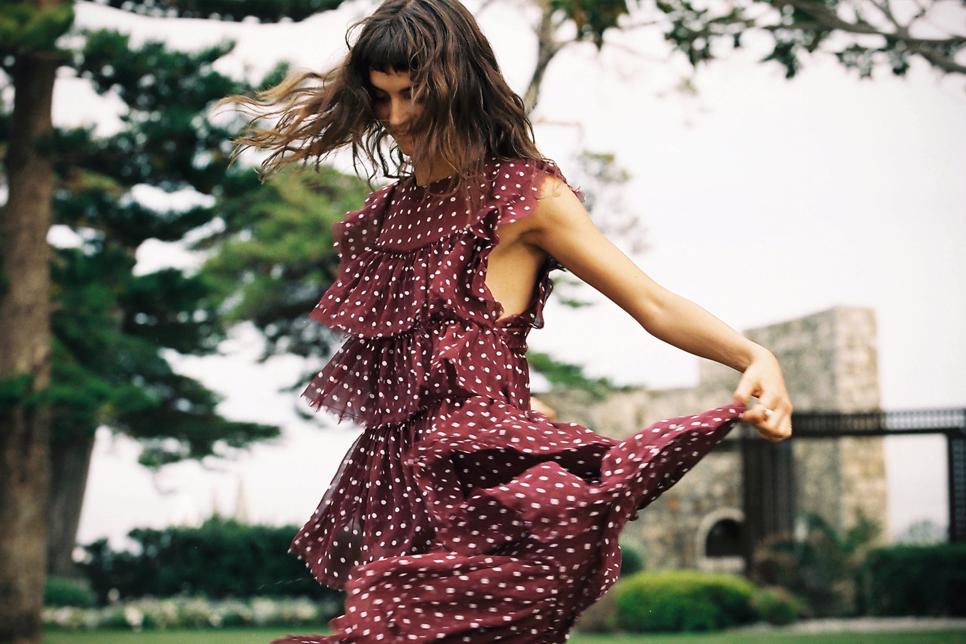 A woman in a maroon polka-dot dress dances in a grassy outdoor setting with trees and a stone structure in the background, creating a fashion editorial.