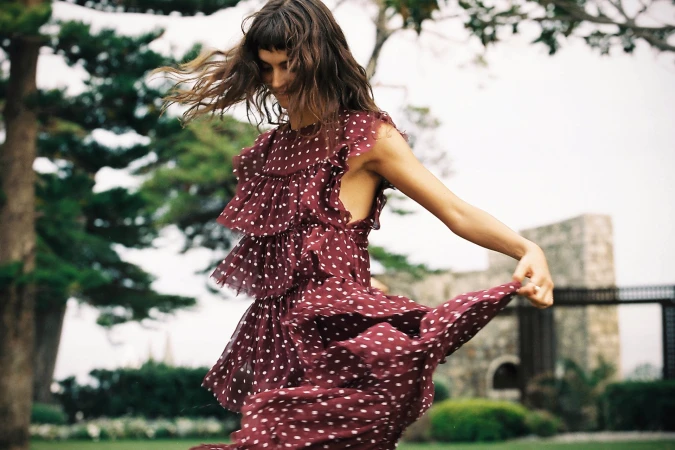 A woman in a maroon polka-dot dress dances in a grassy outdoor setting with trees and a stone structure in the background, creating a fashion editorial.