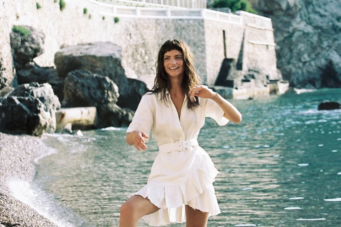 Woman in white belted wrap dress standing by the water with rocky coastline behind her