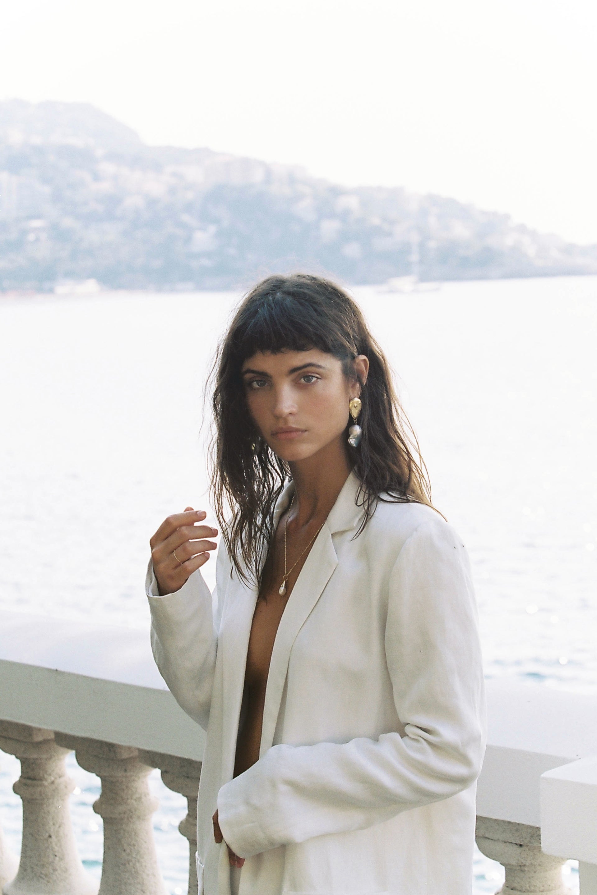 Woman in white linen blazer poses against a bright backdrop of sea and sky, showcasing gold and pearl earrings.