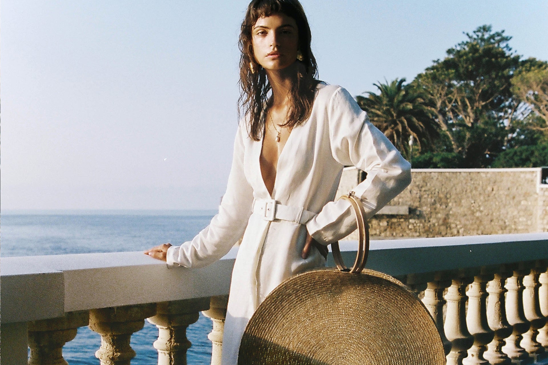 A woman in a white dress stands on a balcony overlooking the ocean, holding a round straw bag, with palm trees and a blue sky in the background.
