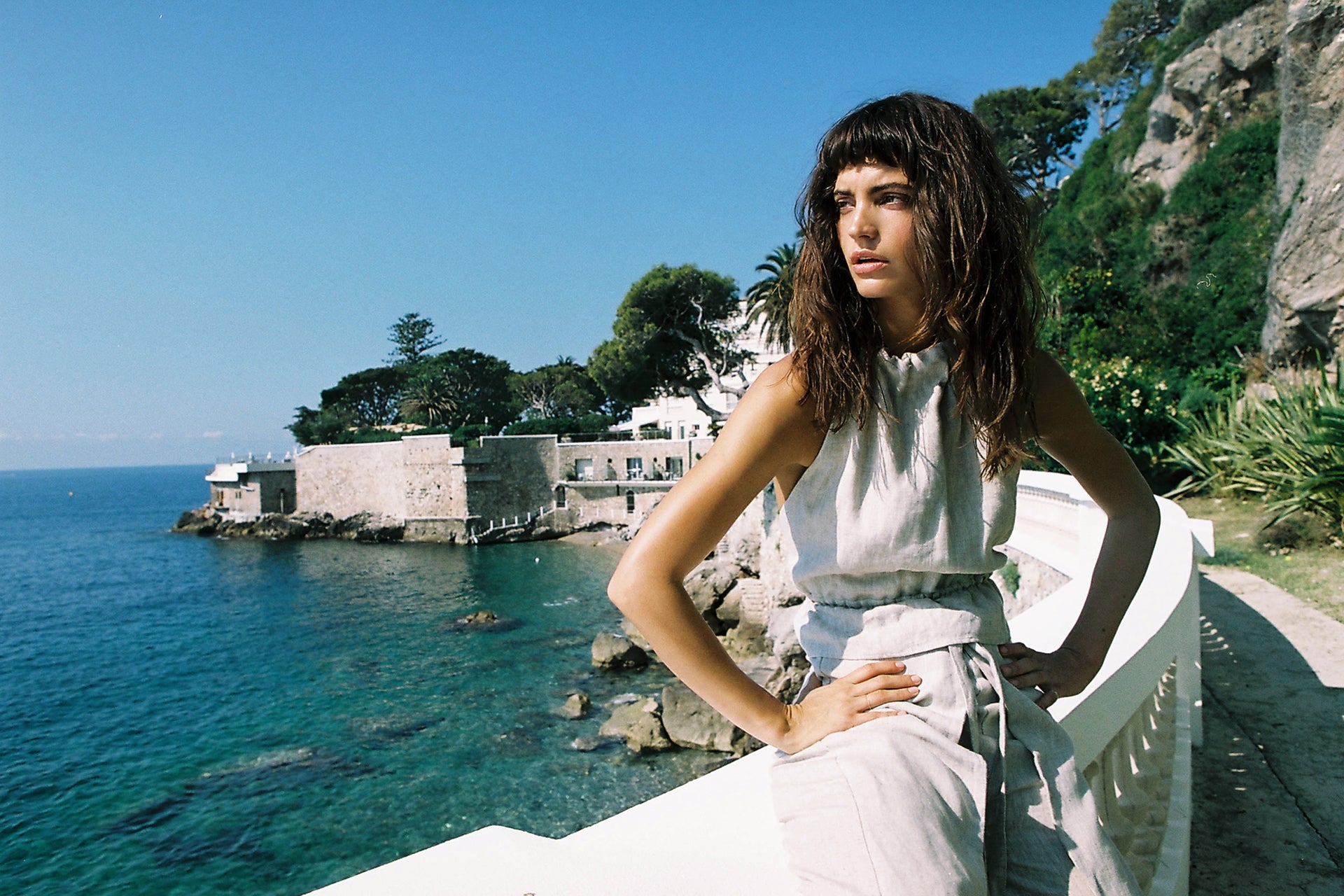 Woman in linen outfit posing on a white stone railing with a coastal view on a bright day with blue sky and turquoise water.