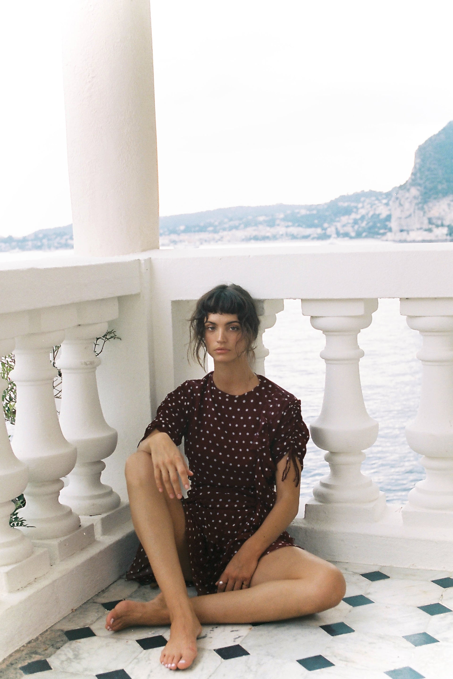 A young adult woman in a dark red polka dot dress poses while sitting cross-legged on a balcony with a sea and mountain view.