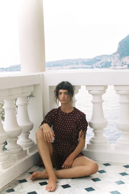 A young adult woman in a dark red polka dot dress poses while sitting cross-legged on a balcony with a sea and mountain view.
