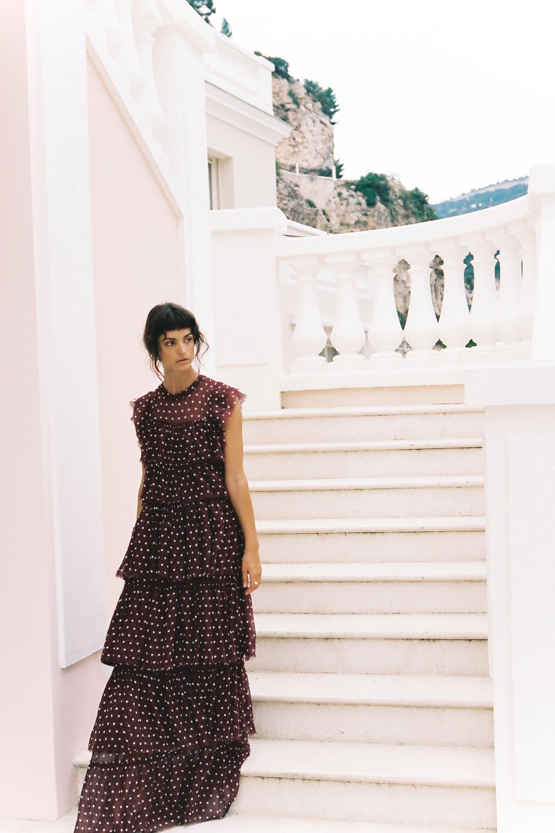 A woman in a tiered dress poses on white stairs with a mountain backdrop, showcasing a fashion editorial style.