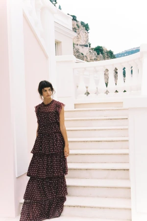 A woman in a tiered dress poses on white stairs with a mountain backdrop, showcasing a fashion editorial style.