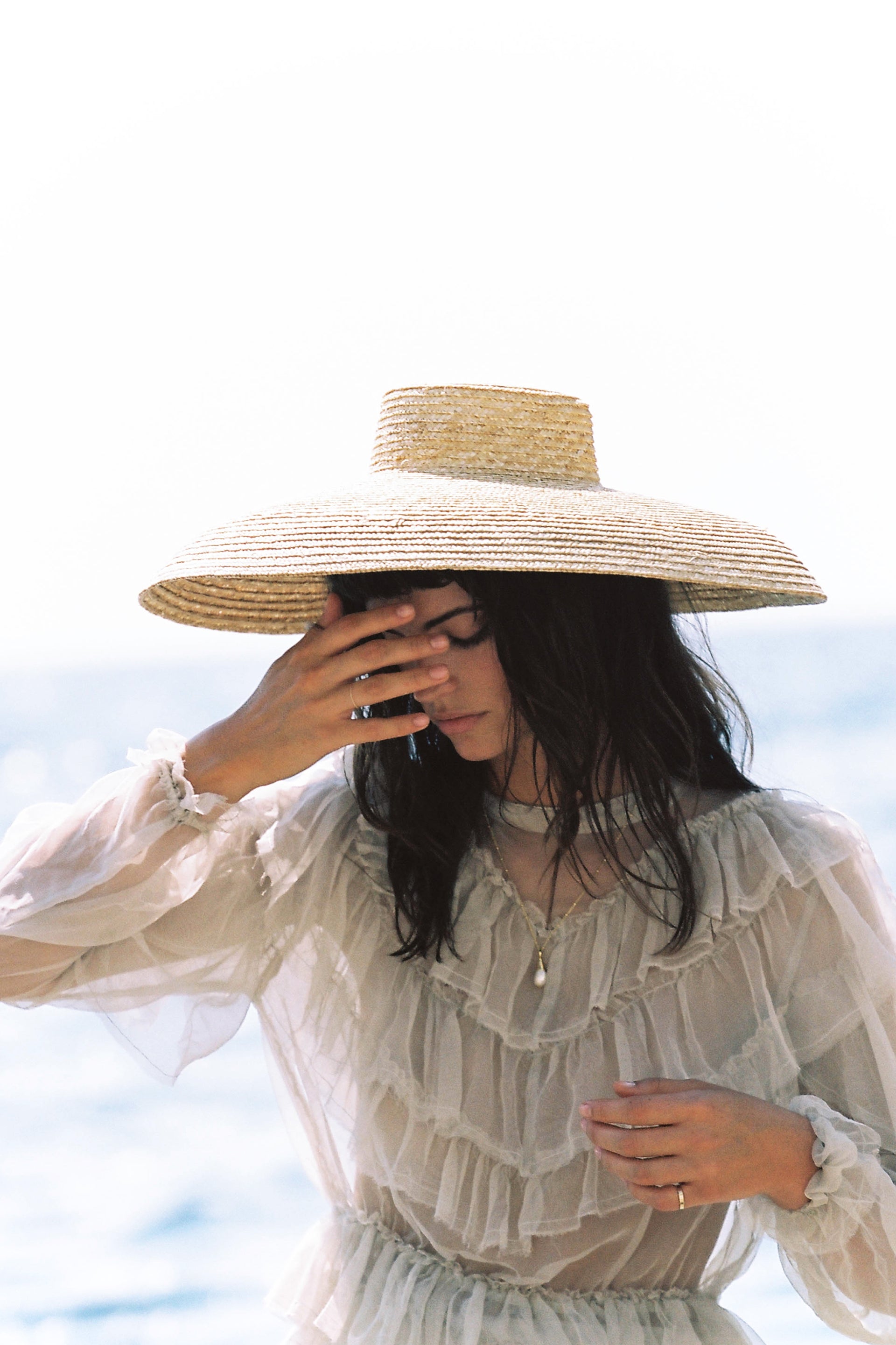 Woman wearing a wide-brimmed straw hat and a sheer tiered ruffled dress posing outdoors by the ocean.