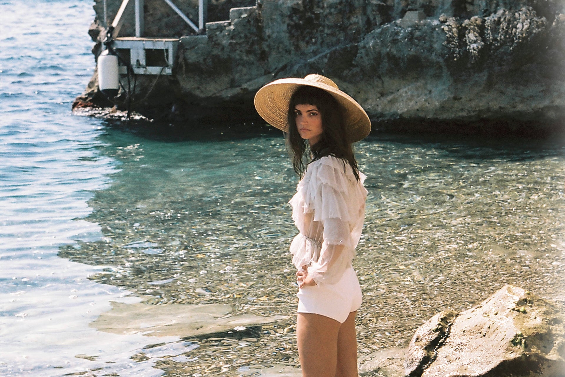 Woman in floppy hat poses near crystal clear ocean water with rocky shoreline, creating a serene, summery fashion editorial.