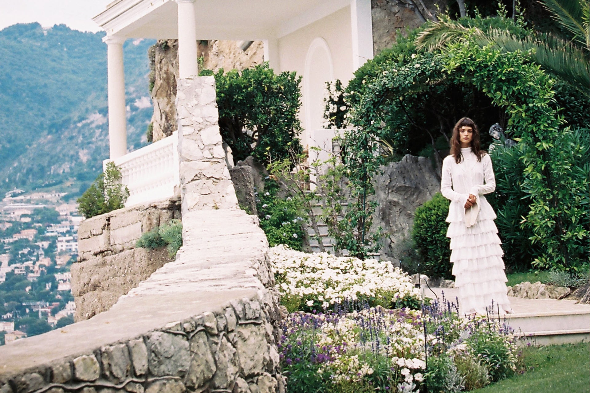A woman poses in a white tiered dress amid lush greenery and blooming flowers, with a stone structure and mountain backdrop.