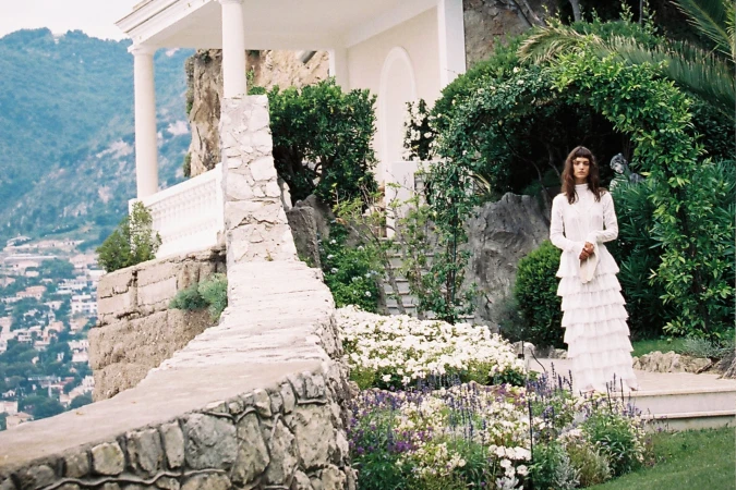A woman poses in a white tiered dress amid lush greenery and blooming flowers, with a stone structure and mountain backdrop.