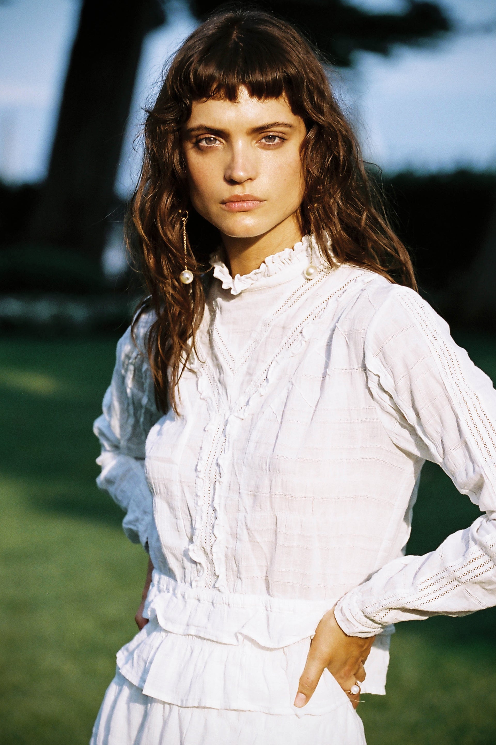 Portrait of a young adult woman with long brown hair, wearing a white blouse, posing outdoors in a sunny, grassy environment.
