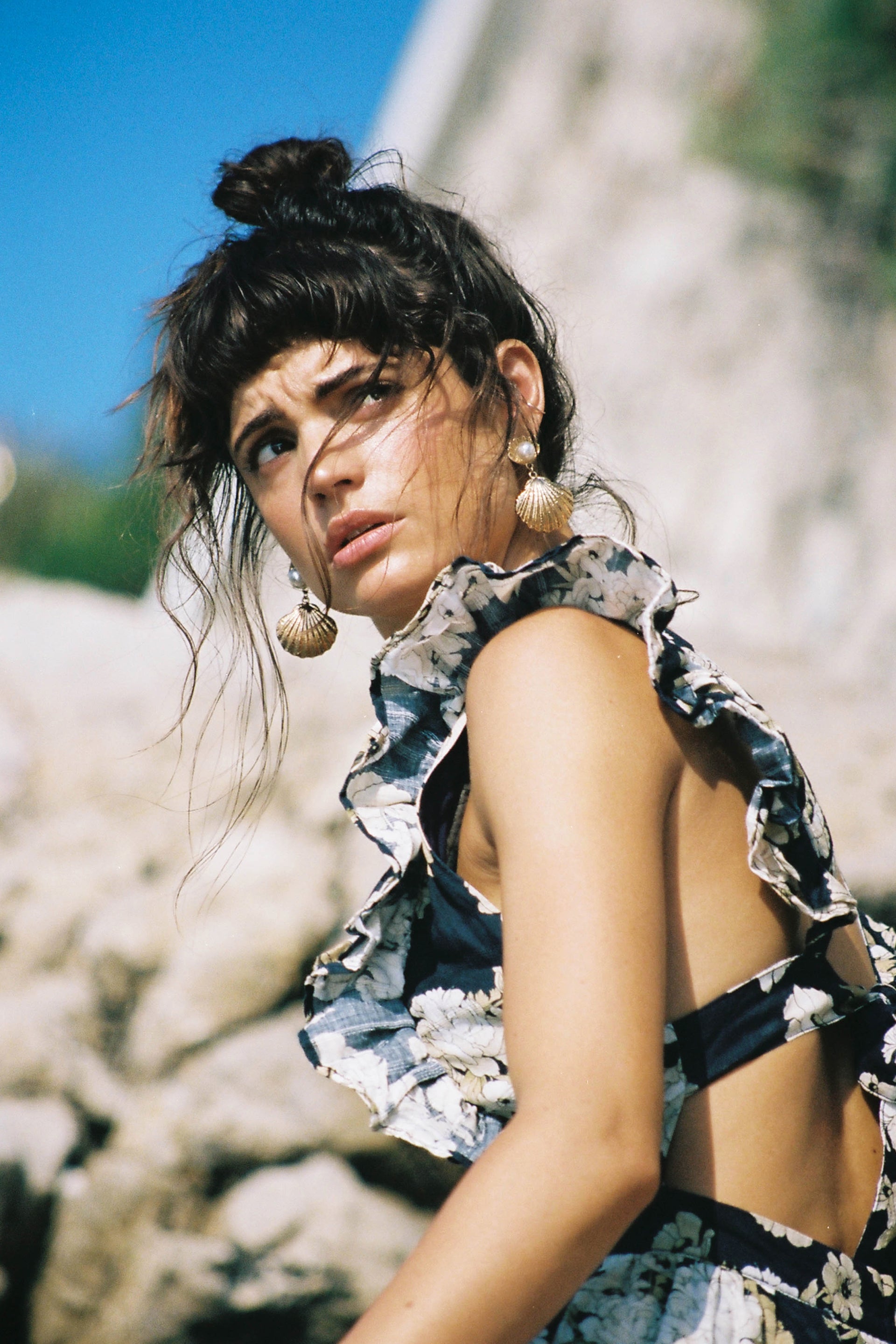 Portrait of a young woman with dark hair in a bun wearing seashell earrings and a floral print top against a stone background.