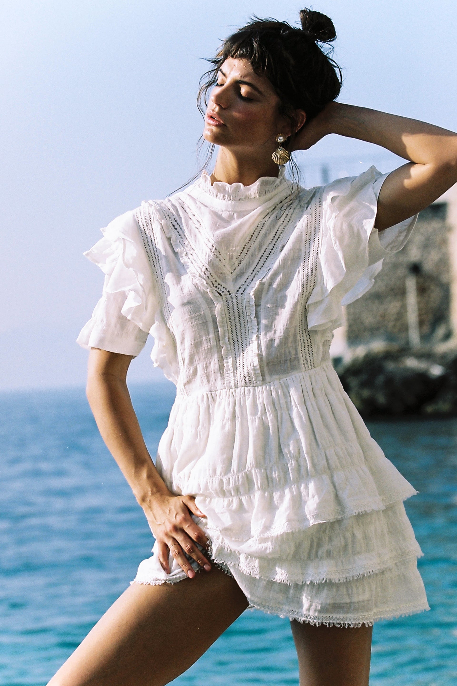 Woman in a white dress poses against a sunny outdoor backdrop, ocean visible in the distance, creating a serene fashion portrait.