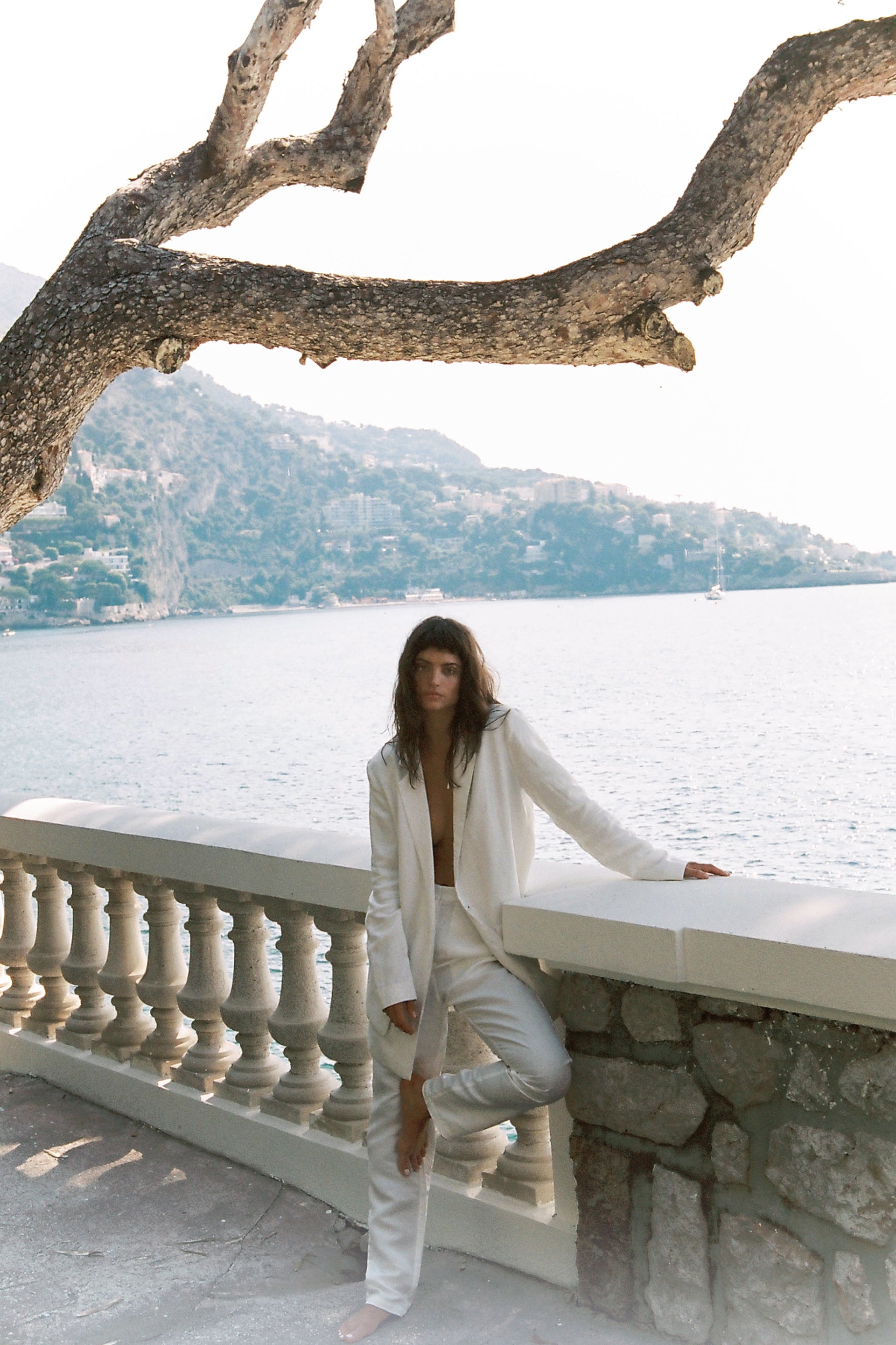 Woman in white suit posing on a stone ledge overlooking the sea with mountains and a tree branch in the background on a bright day.