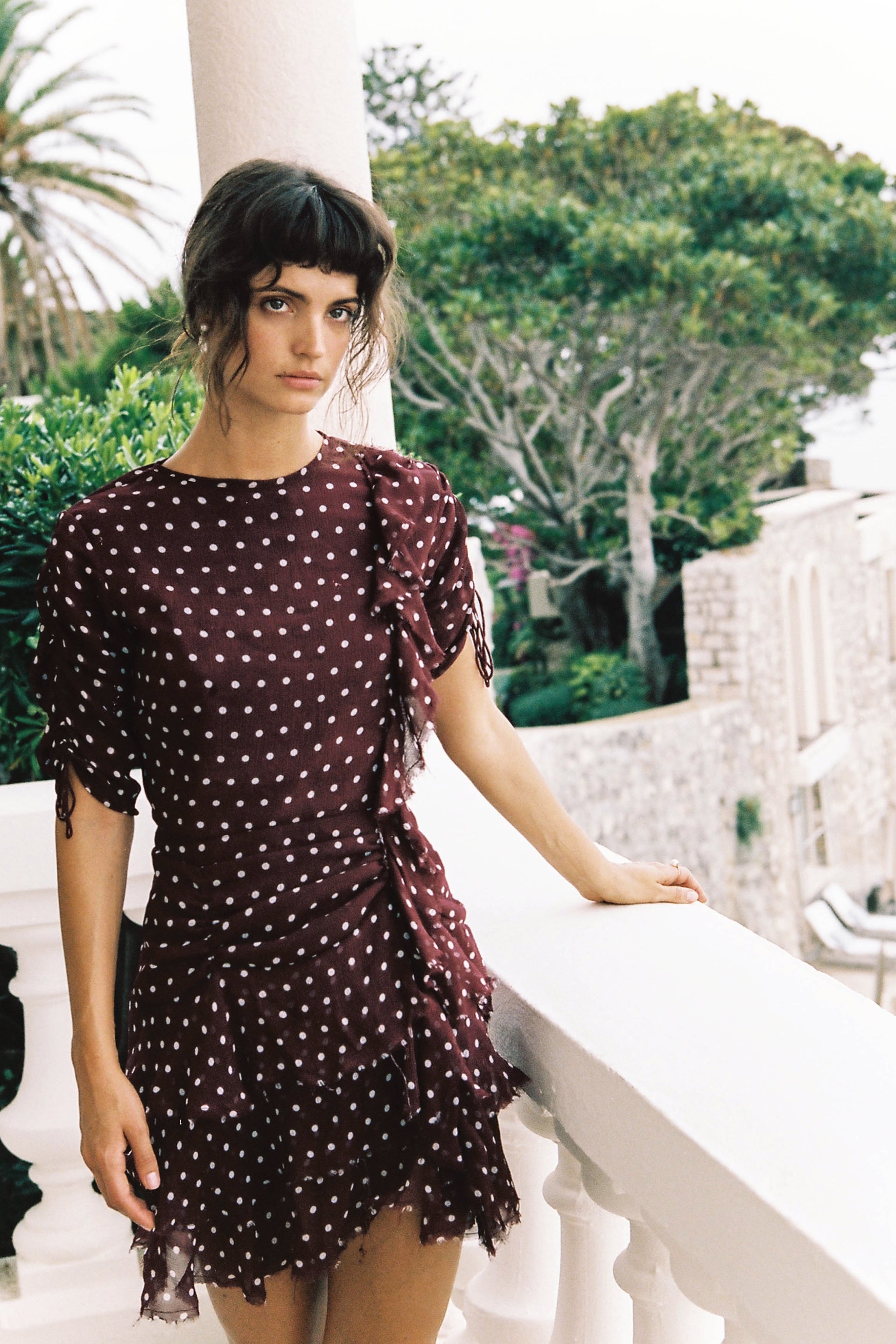 Woman in a dark burgundy polka dot mini dress posing on a white balcony with lush green trees in the background