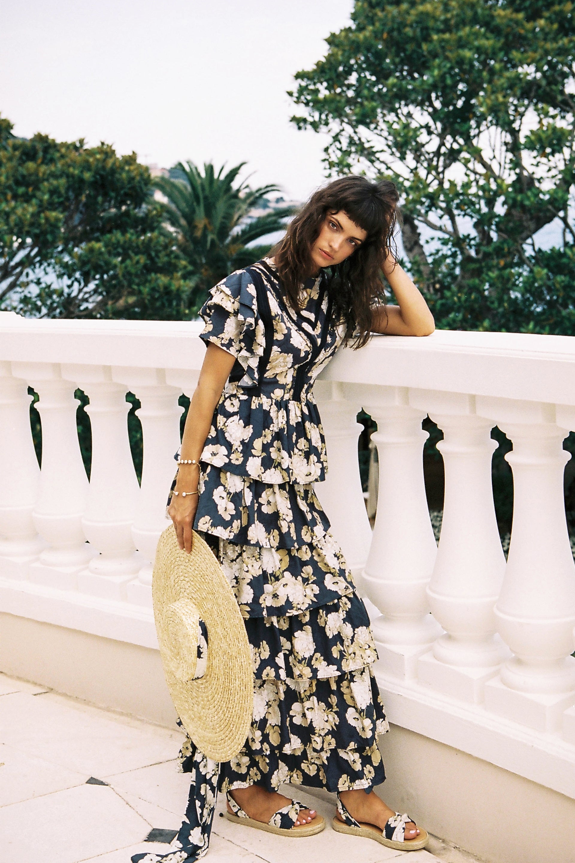 Fashion model wearing a floral print dress with a straw hat and sandals poses on a balcony with a green landscape background.