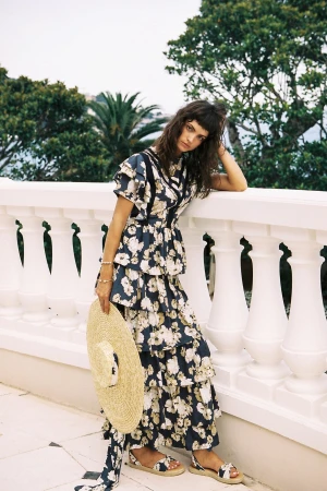Fashion model wearing a floral print dress with a straw hat and sandals poses on a balcony with a green landscape background.
