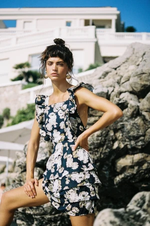 Model wearing a dark floral print minidress standing against rocky terrain with a white building in the background.