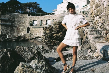 Model standing on rocky coastal shore wearing a white ruffled mini dress and tied sandals for Sir The Label Spring by Justin Woo