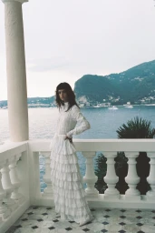 Model in a long white ruffled dress posing on a white balustrade balcony overlooking a coastal body of water and mountains.