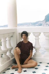 Woman in a burgundy polka dot dress seated on a tiled balcony overlooking the ocean by Justin Woo