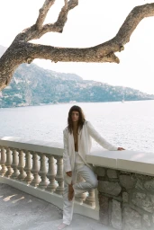 Woman in white suit leaning against a stone balustrade overlooking a coastal town and sea under a large tree branch