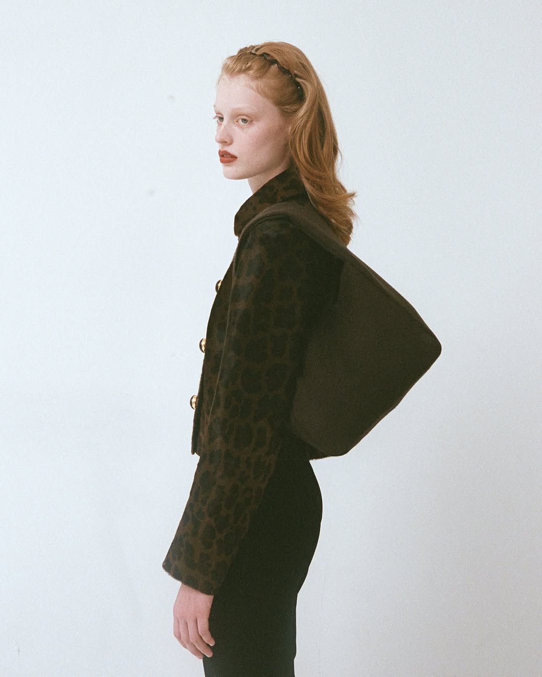 A woman with auburn hair poses in a patterned brown jacket with a matching bag against a white background in a fashion shot.