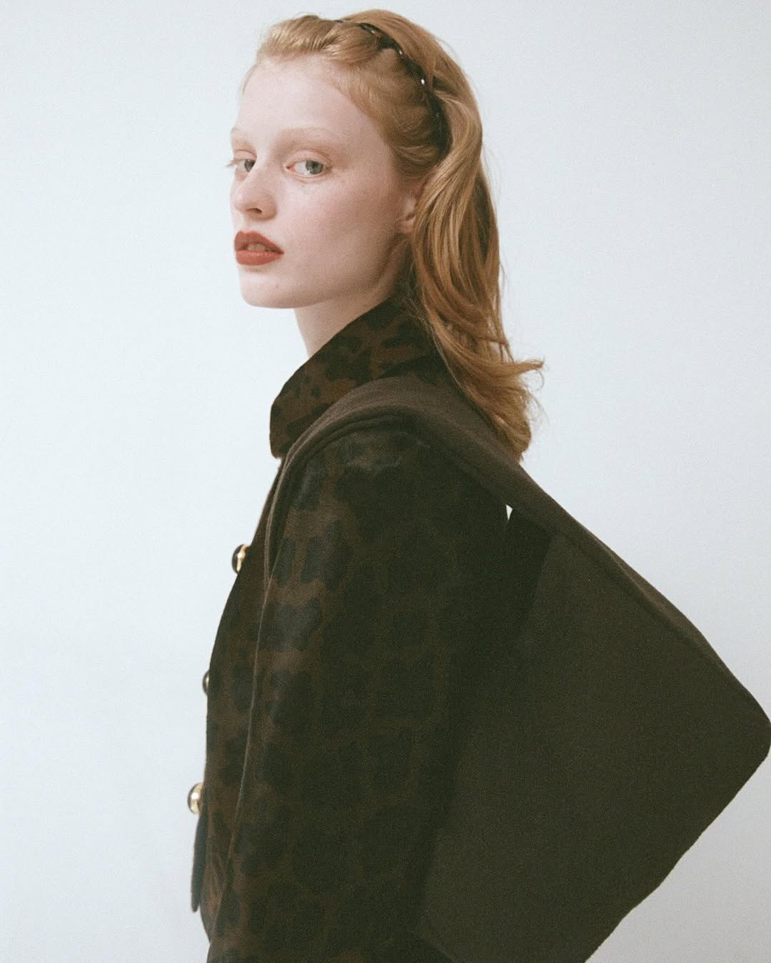 Portrait of a red-haired woman with a dark brown, patterned cape and gold buttons against a white background in a studio setting.