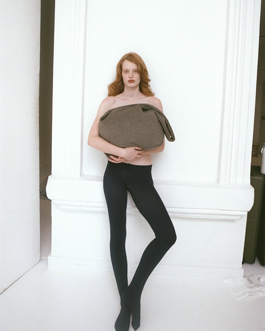 Fashion shot of a redhead model holding a large bag in a studio setting, wearing black tights and posing against a white column.