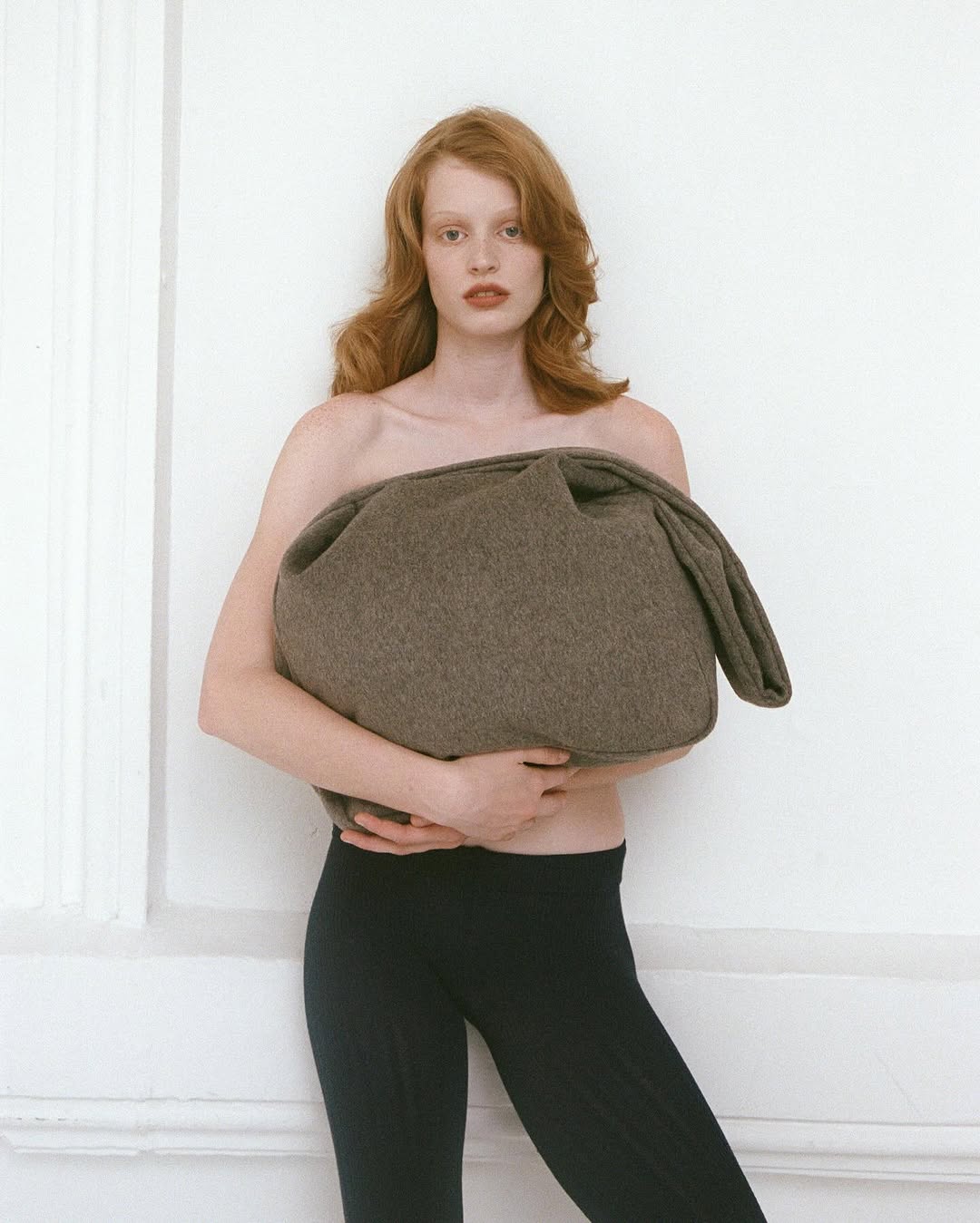 Red-haired model holding a large brown wool bag against a plain white wall