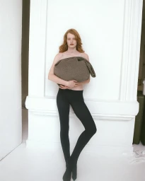 Red-haired model wearing black tights holding a large chocolate wool pouch against a white wall.