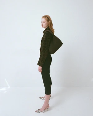 Model standing against a white backdrop wearing a patterned dark jacket, black trousers, and carrying a wool shoulder bag.