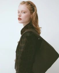 Red-haired model wearing a dark chocolate wool garment holding a matching handbag against a plain white background