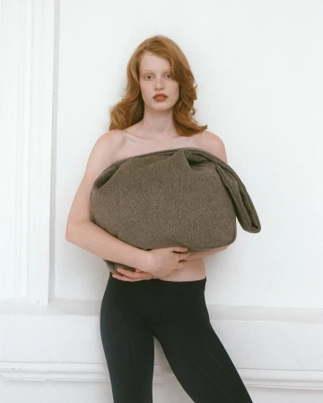 Red-haired model holding a large brown wool bag against a plain white wall